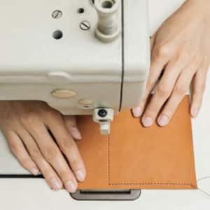 Person using a sewing machine to stitch two pieces of tan leather fabric.