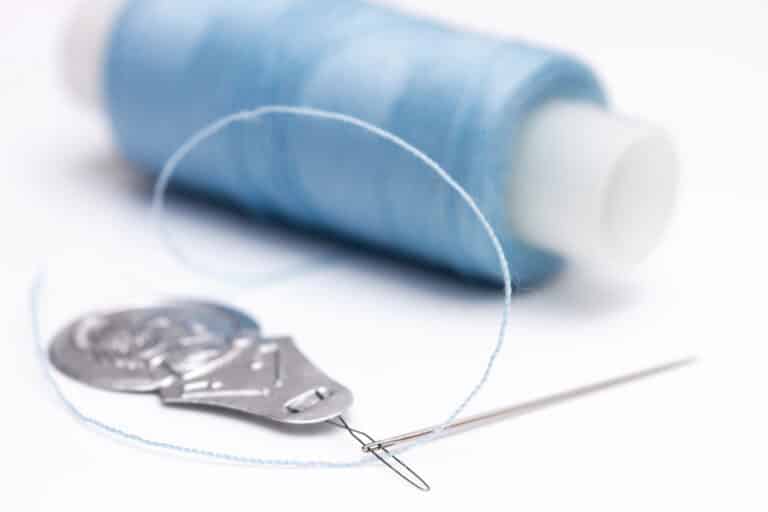 A close-up of a threaded needle with a needle threader, spool of blue thread, and a small pair of scissors against a white background.